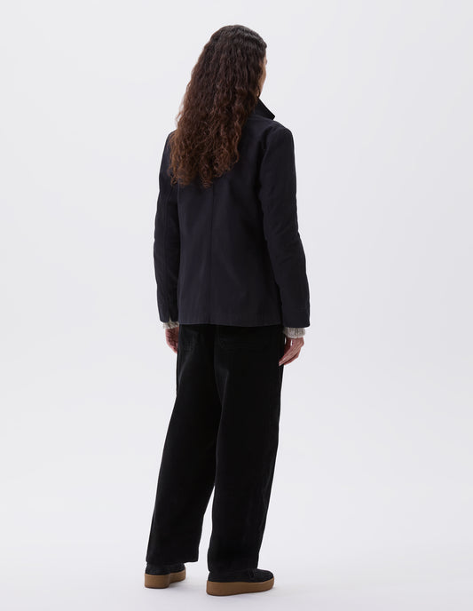Full body image, back view of model wearing indigo twill single breasted blazer, with fold back split cuff and curved hem. Styled over beige jumper, black trousers and black suede trainers.