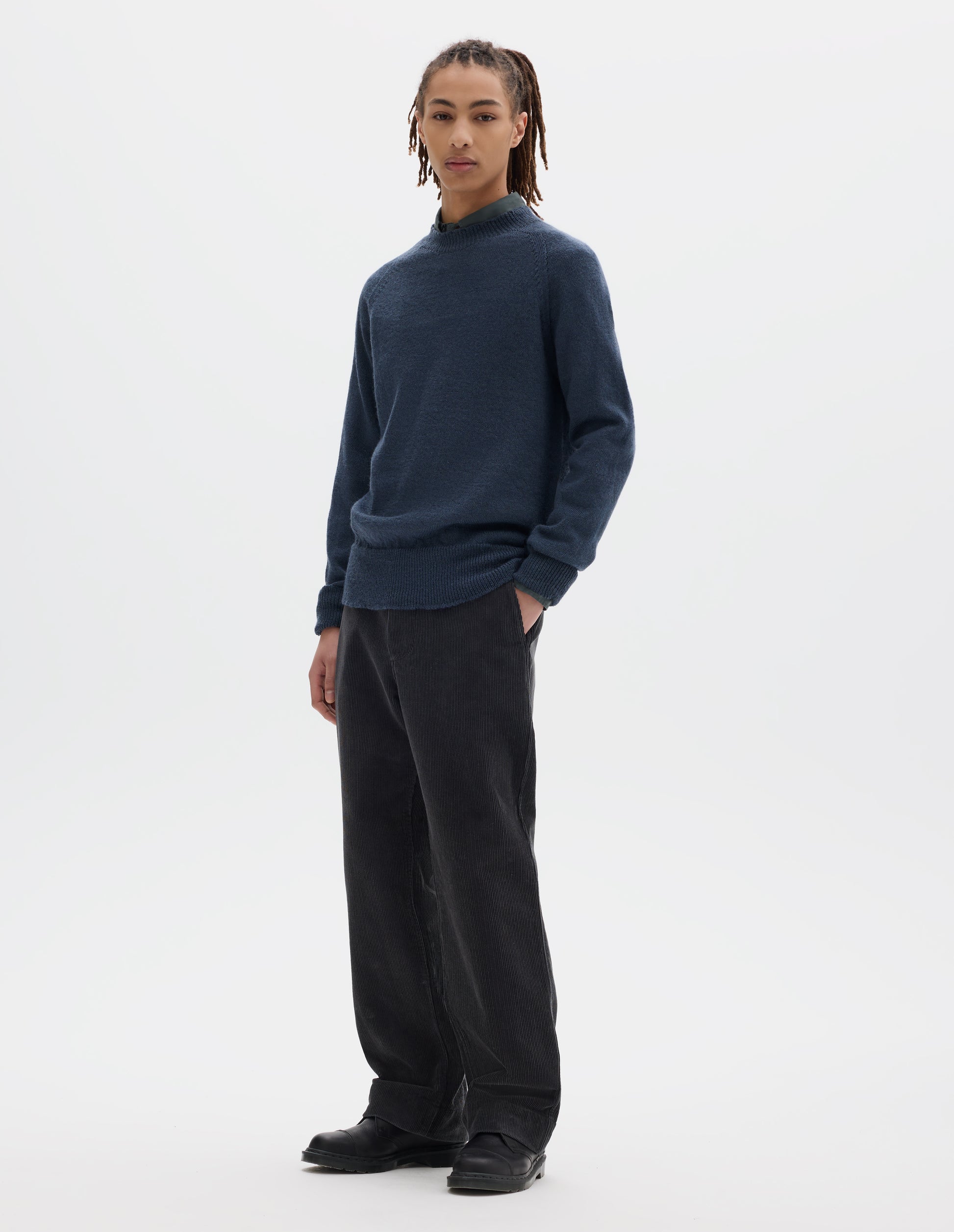 Full body image, front view of model wearing a blue crew neck wool jumper with raglan sleeves and rib collar, cuffs and hem. Worn over a blue shirt and styled with dark grey striaght leg trousers and black leather shoes.