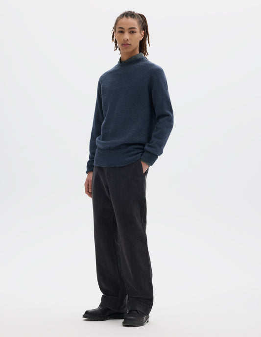 Full body image, front view of model wearing a blue crew neck wool jumper with raglan sleeves and rib collar, cuffs and hem. Worn over a blue shirt and styled with dark grey striaght leg trousers and black leather shoes.