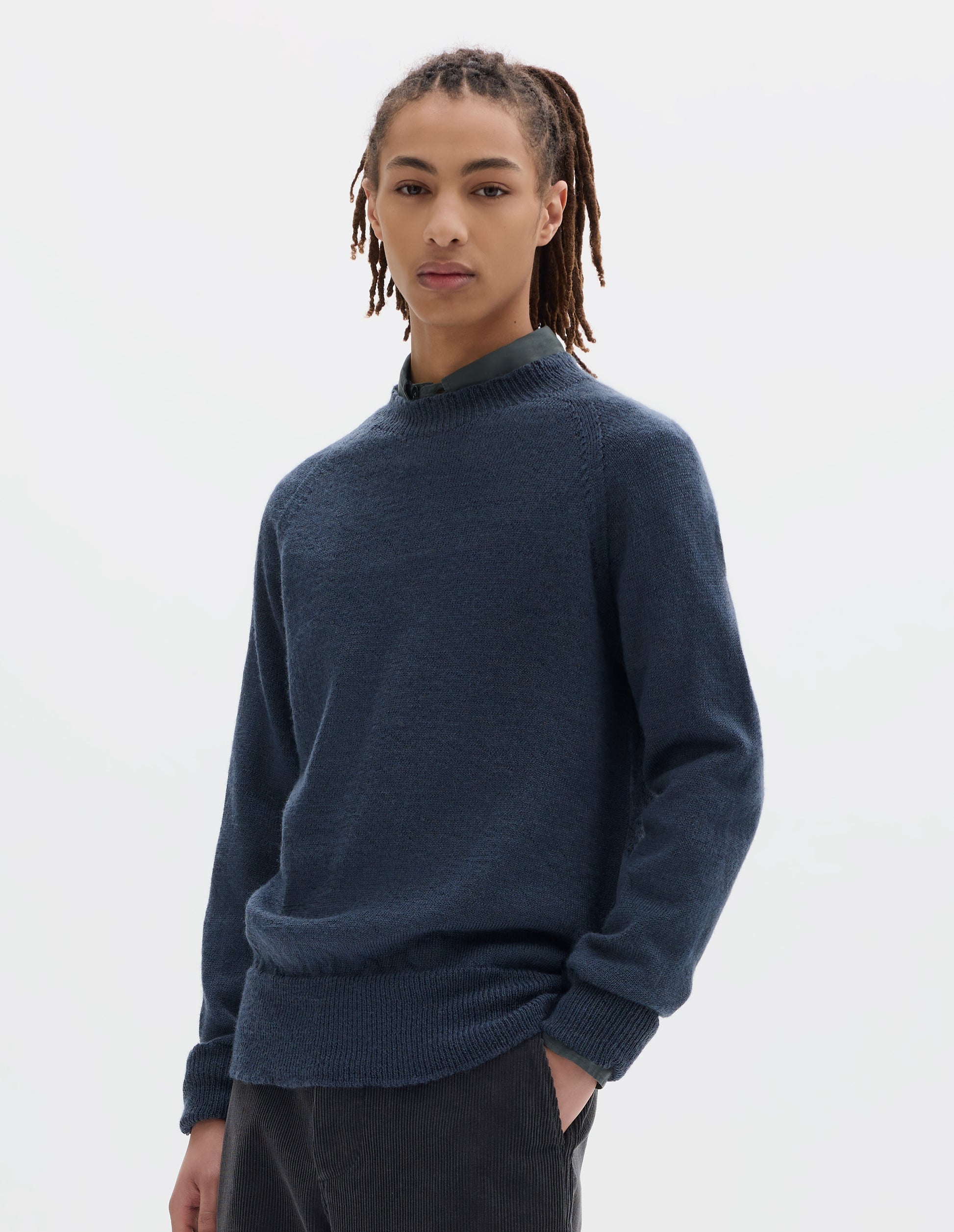 Cropped body image, front view of model wearing a blue crew neck wool jumper with raglan sleeves and rib collar, cuffs and hem. Worn over a blue shirt and styled with dark grey trousers.