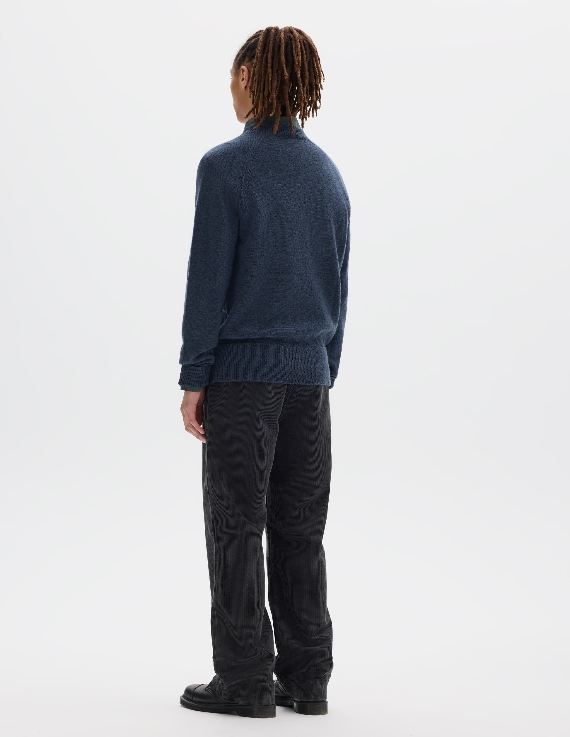 Full body image, back view of model wearing a blue crew neck wool jumper with raglan sleeves and rib collar, cuffs and hem. Worn over a blue shirt and styled with dark grey striaght leg trousers and black leather shoes.