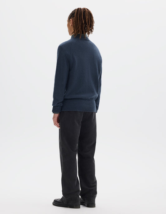 Full body image, back view of model wearing a blue crew neck wool jumper with raglan sleeves and rib collar, cuffs and hem. Worn over a blue shirt and styled with dark grey striaght leg trousers and black leather shoes.