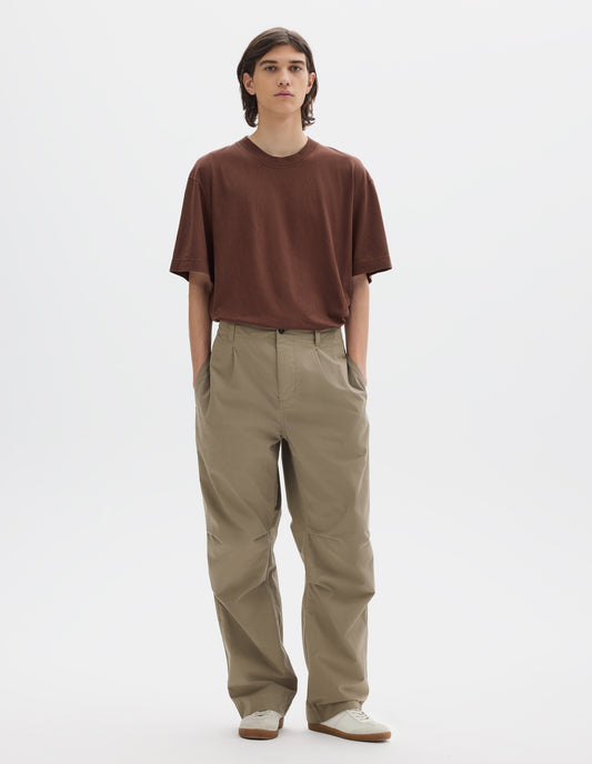 Full body image, front view of model wearing a brown short sleeve T shirt, greyish beige cotton trousers with side seam pockets, single front pleat and pleated knee detailing. Styled with off white suede trainers.