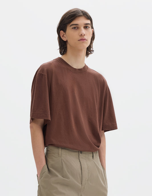 Cropped body image, front view of model wearing a brown short sleeve cotton linen t shirt, tucked into beige pleated cotton trousers with side seam pockets.