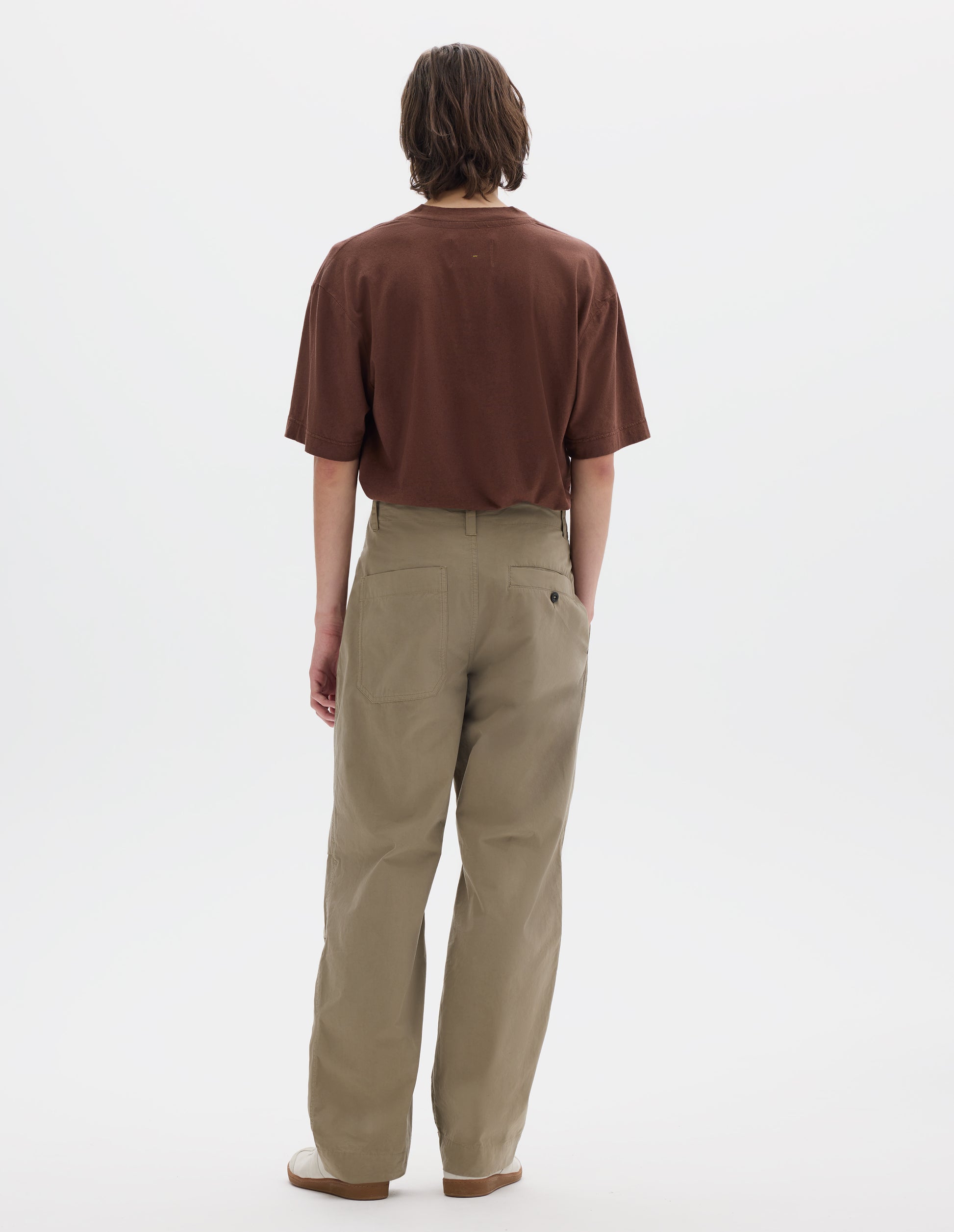 Full body image, back view of model wearing a brown short sleeve cotton linen t shirt, tucked into beige cotton trousers with side seam pockets and two back pockets, one is a patch pocket and one a button through welt pocket. Styled with off white suede trainers. 