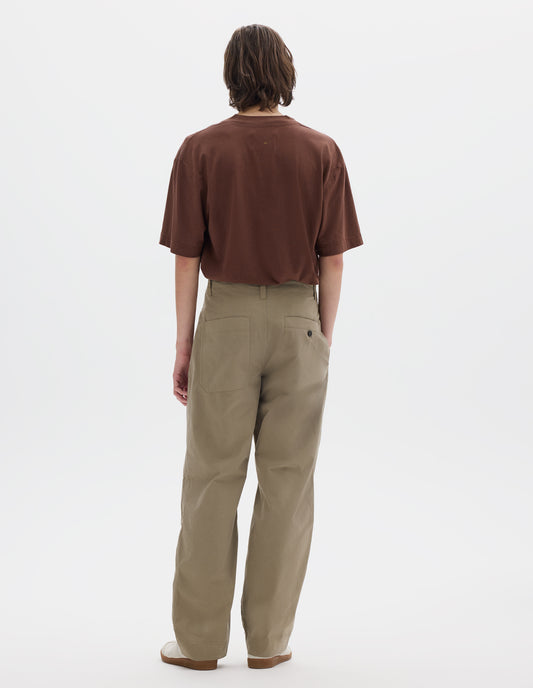 Full body image, back view of model wearing a brown short sleeve cotton linen t shirt, tucked into beige cotton trousers with side seam pockets and two back pockets, one is a patch pocket and one a button through welt pocket. Styled with off white suede trainers. 