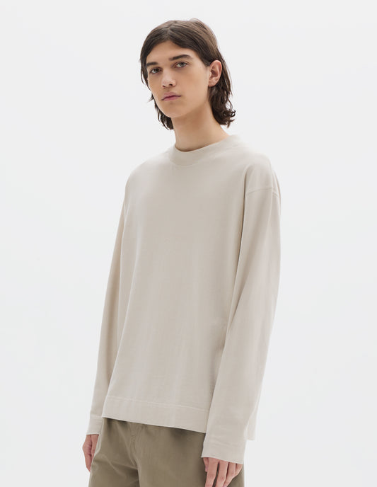 Cropped body image, front view of model wearing an off white long sleeve, flatlock stitching, a deep ribbed collar and ribbed triangle insert on the shoulder. Styled with faded khaki green trousers.