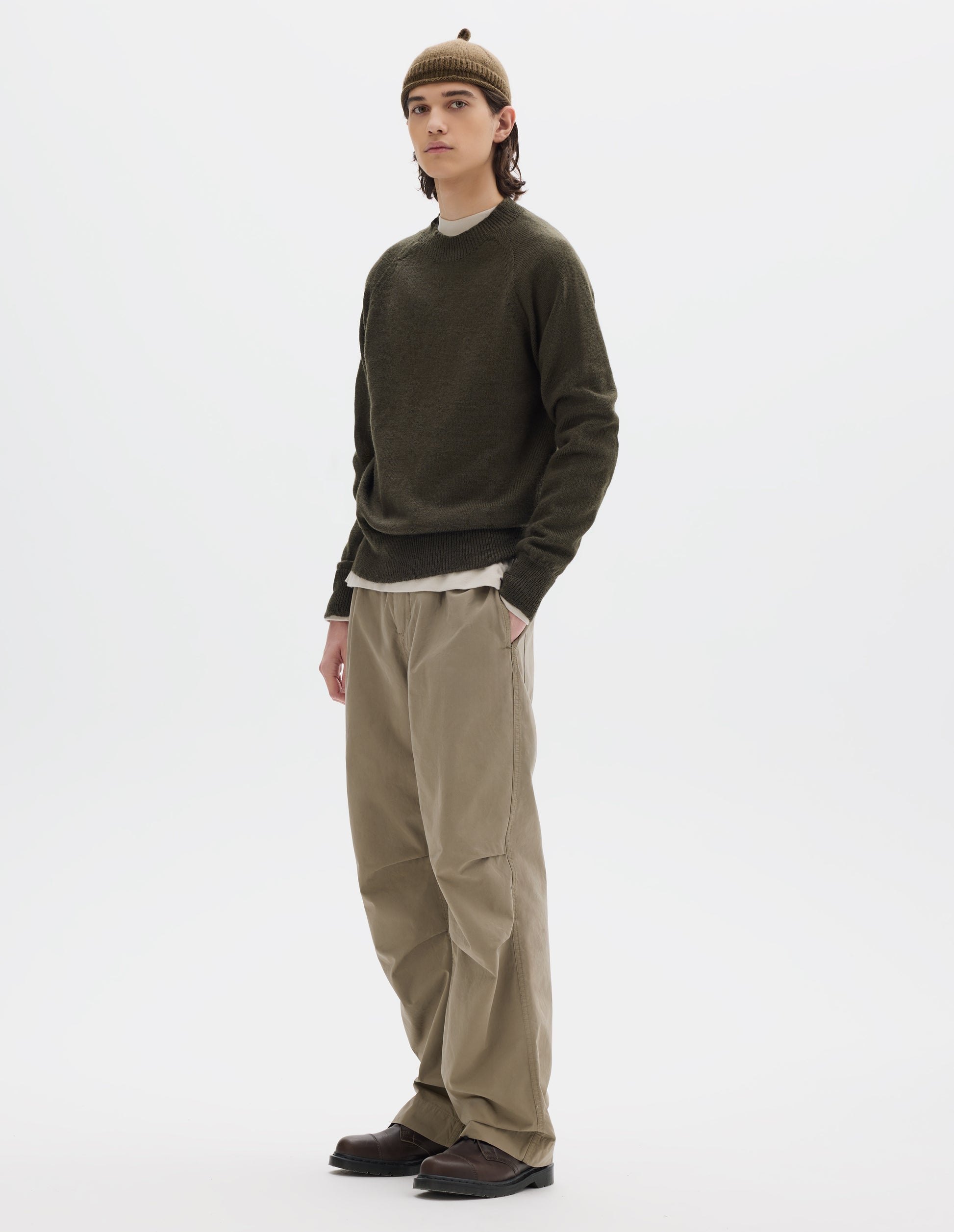 Full body image, front view of model wearing a green-brown crew neck wool jumper with raglan sleeves and rib collar, cuffs and hem. Worn over an off white jersey and styled with a brown wool beanie with knitted loop detailing on top, beige trousers and brown leather shoes.