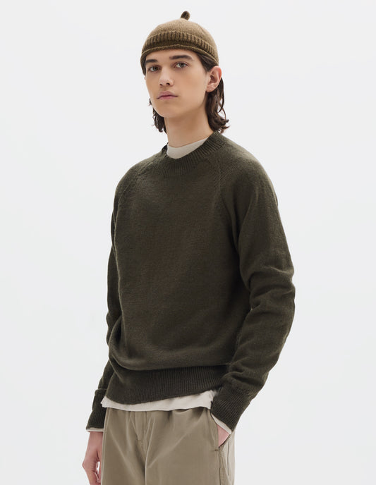 Cropped body image, front view of model wearing a green-brown crew neck wool jumper with raglan sleeves and rib collar, cuffs and hem. Worn over an off white jersey and styled with a brown wool beanie with knitted loop detailing on top and beige trousers.