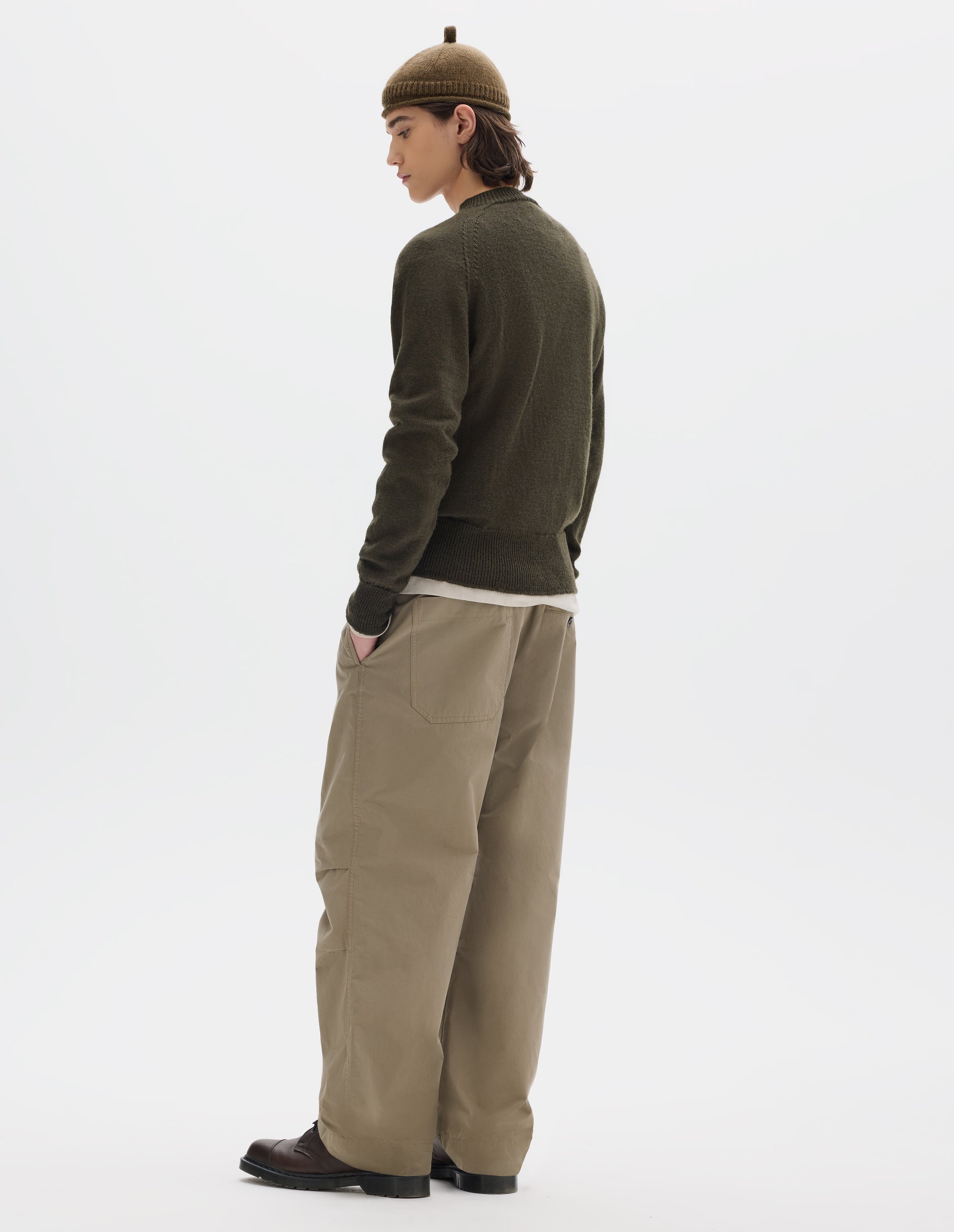 Full body image, back view of model wearing a green-brown crew neck wool jumper with raglan sleeves and rib collar, cuffs and hem. Worn over an off white jersey and styled with a brown wool beanie with knitted loop detailing on top, beige trousers and brown leather shoes.