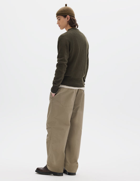 Full body image, back view of model wearing a green-brown crew neck wool jumper with raglan sleeves and rib collar, cuffs and hem. Worn over an off white jersey and styled with a brown wool beanie with knitted loop detailing on top, beige trousers and brown leather shoes.
