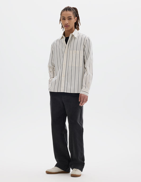 Full body image, front view of model wearing an off white and dark blue stripe cotton shirt, with light buttons and button-through chest patch pocket. Styled over a black T shirt and dark grey cord trousers and pale beige trainers.