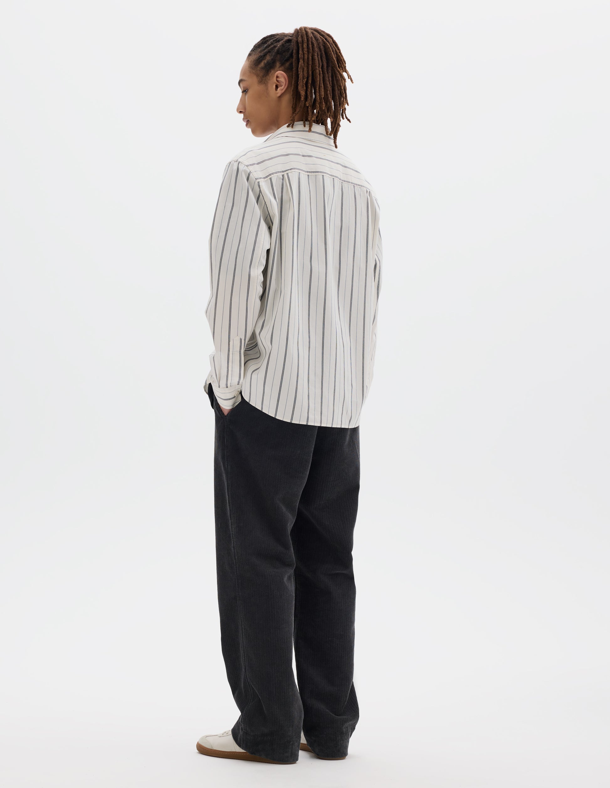 Full body image, back view of model wearing an off white and dark blue stripe cotton shirt, with two pleats on back yoke. Styled with dark grey cord trousers and pale beige trainers.
