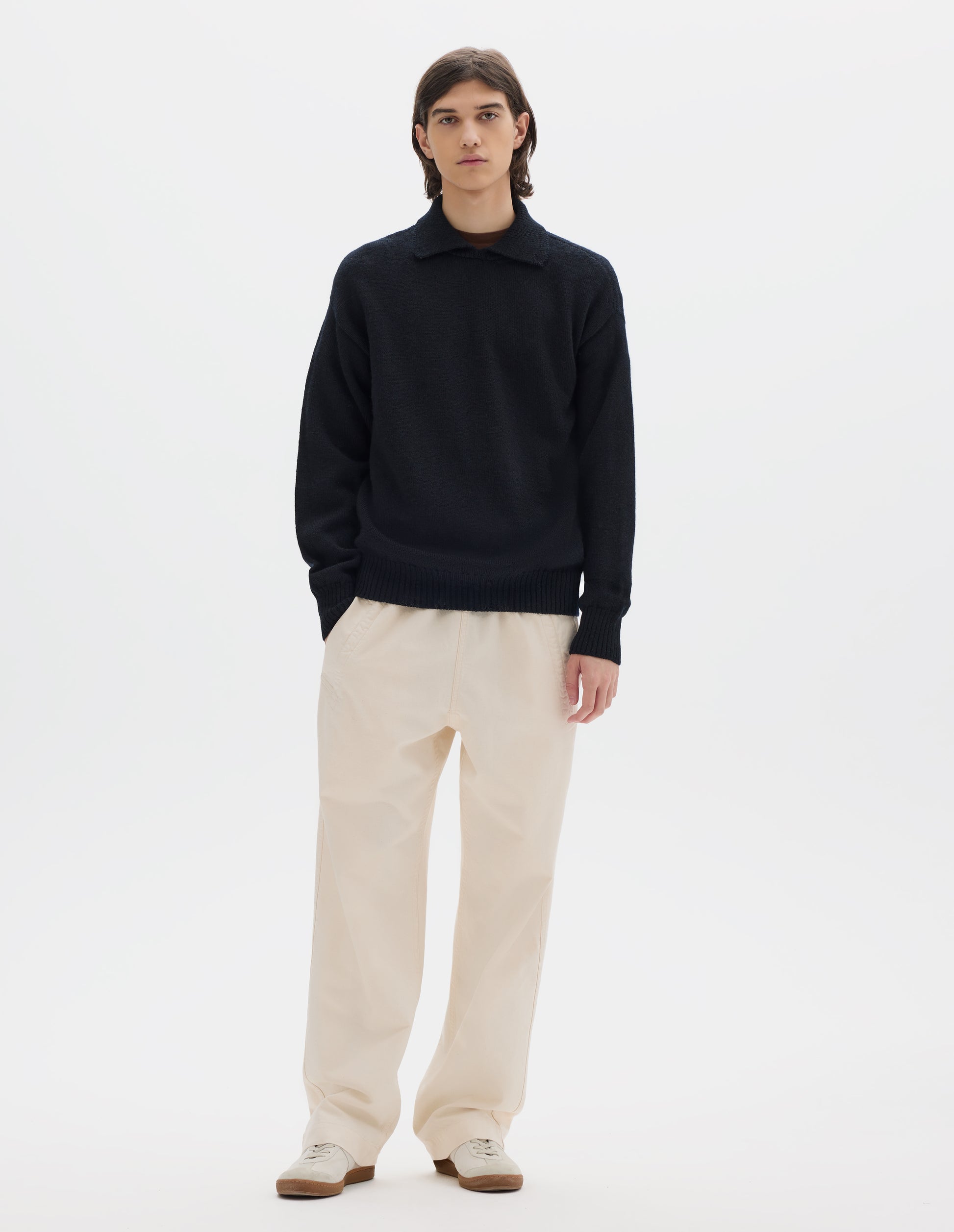 Full body image, front view of model wearing a black wool collared sweater  with a wide rib linked on collar, plain knit body, and deeper ribbed cuffs. Styled with off white jogger style trousers and white suede trainers.