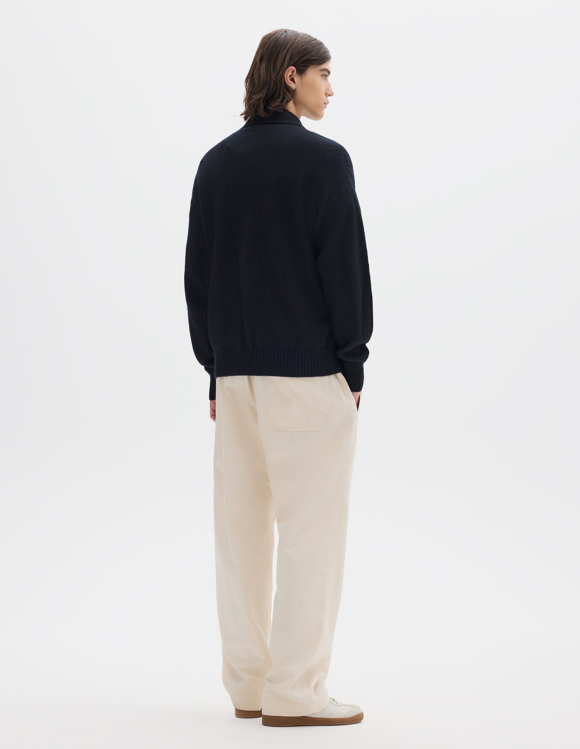 Full body image, back view of model wearing a black wool collared sweater  with a wide rib linked on collar, plain knit body, and deeper ribbed cuffs. Styled with off white jogger style trousers and white suede trainers.