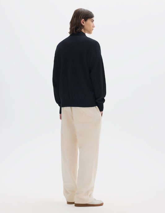 Full body image, back view of model wearing a black wool collared sweater  with a wide rib linked on collar, plain knit body, and deeper ribbed cuffs. Styled with off white jogger style trousers and white suede trainers.