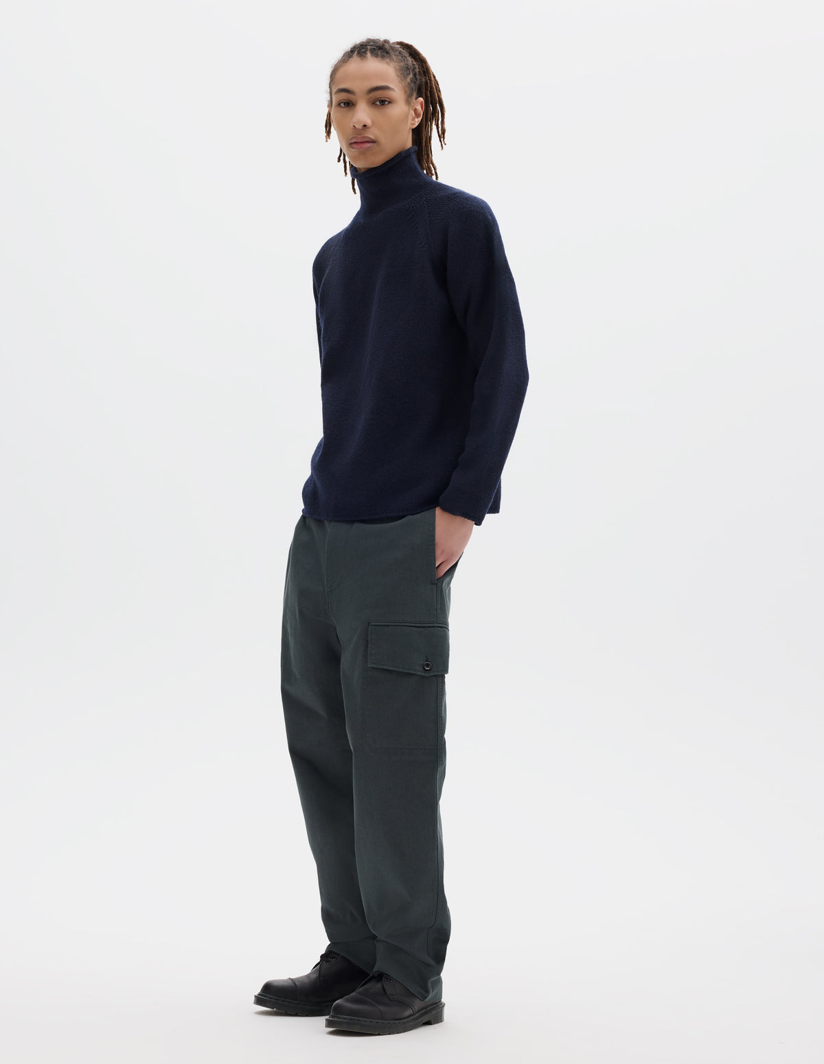 Full body image, front view of model wearing a dark blue high neck wool sweater with raglan sleeves, rolled cuffs and hem. Styled with dark grey cotton trousers which feature a cargo pocket, and black leather shoes.