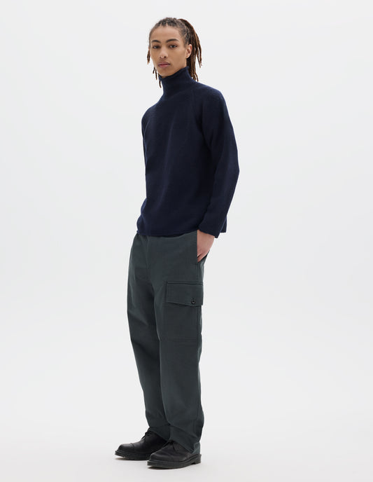 Full body image, front view of model wearing a dark blue high neck wool sweater with raglan sleeves, rolled cuffs and hem. Styled with dark grey cotton trousers which feature a cargo pocket, and black leather shoes.
