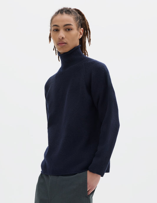 Cropped body image, front view of model wearing a dark blue high neck wool sweater with raglan sleeves, rolled cuffs and hem. Styled with dark grey cotton trousers.
