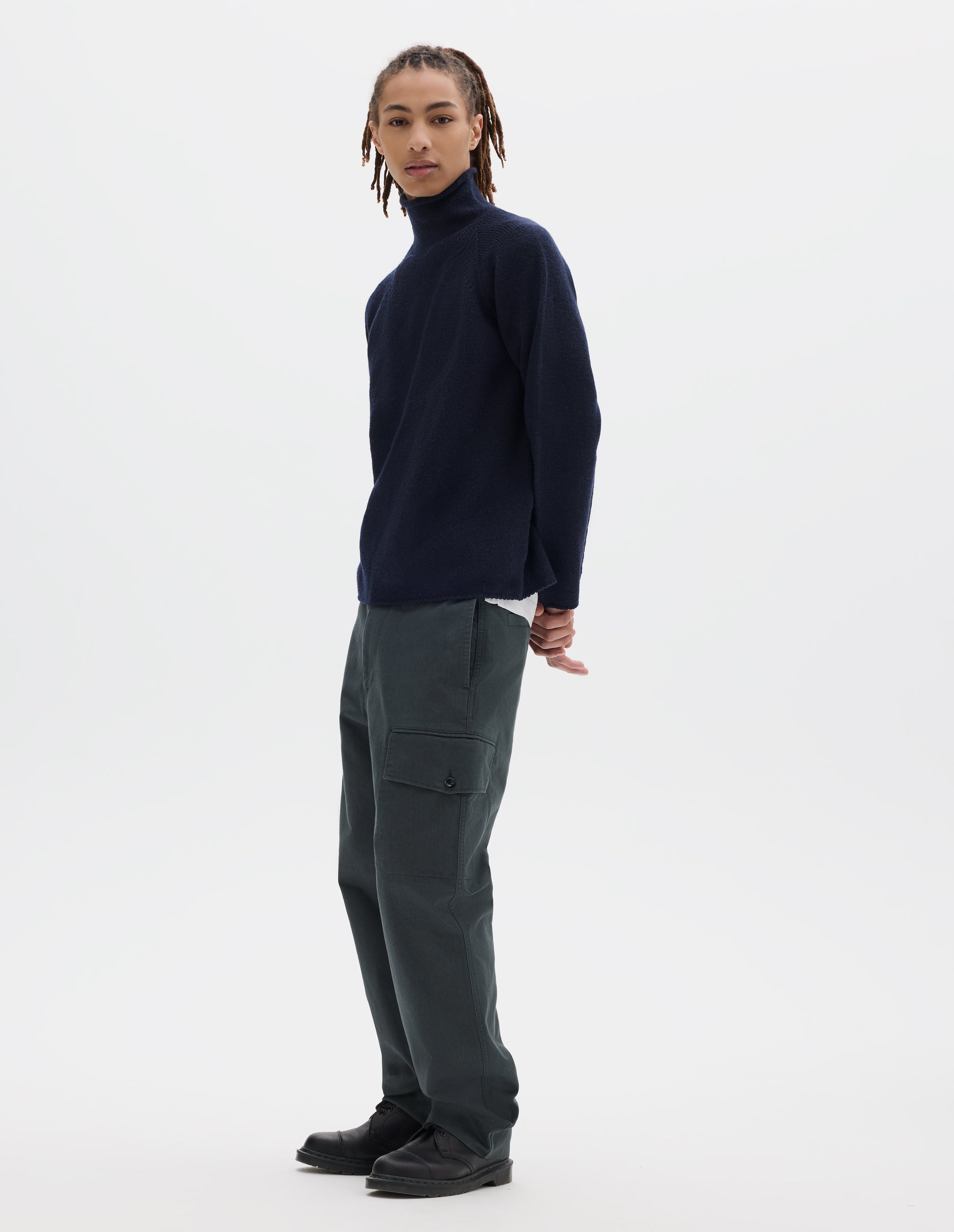 Full body image, front view of model wearing a navy high neck wool jumper, worn over a white T shirt and styled with dark grey cargo trousers with a buttoned pocket and black leather shoes.