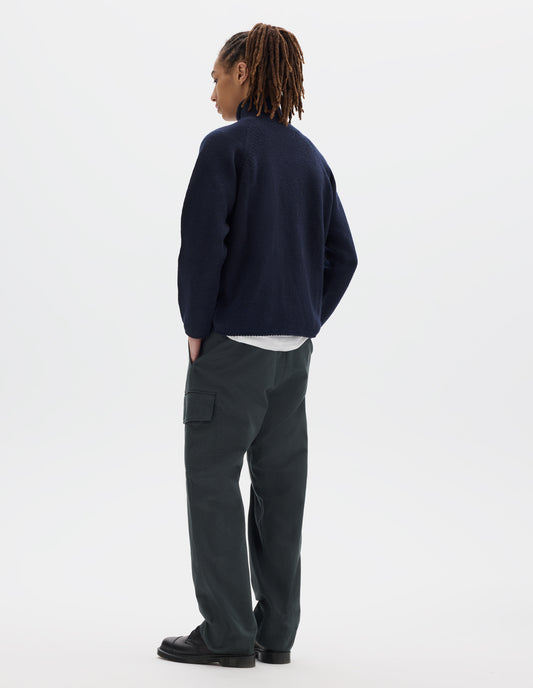 Full body image, back view of model wearing a dark blue high neck wool sweater with raglan sleeves, rolled cuffs and hem. Styled over a white jersey, with dark grey cotton trousers which feature a cargo pocket, and black leather shoes.