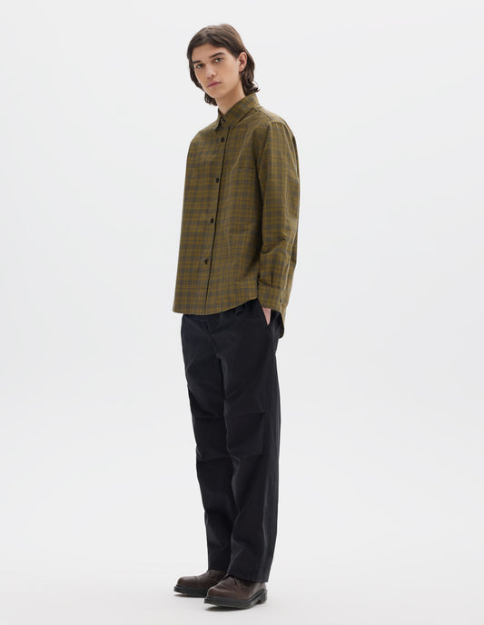 Full body image, front view of model wearing a green check shirt with a raised placket, chest pocket and a curved hem. Worn with black cotton trousers with pleated knee detailing. Styled with brown leather shoes.
