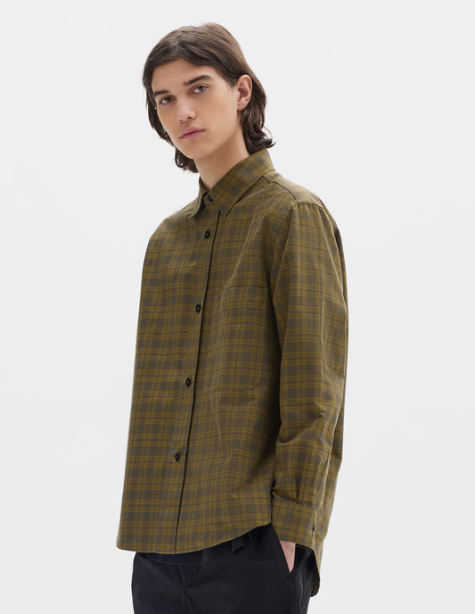 Cropped body image, front view of model wearing a green check shirt with a raised placket, chest pocket and curved hem. Worn with black cotton trousers.