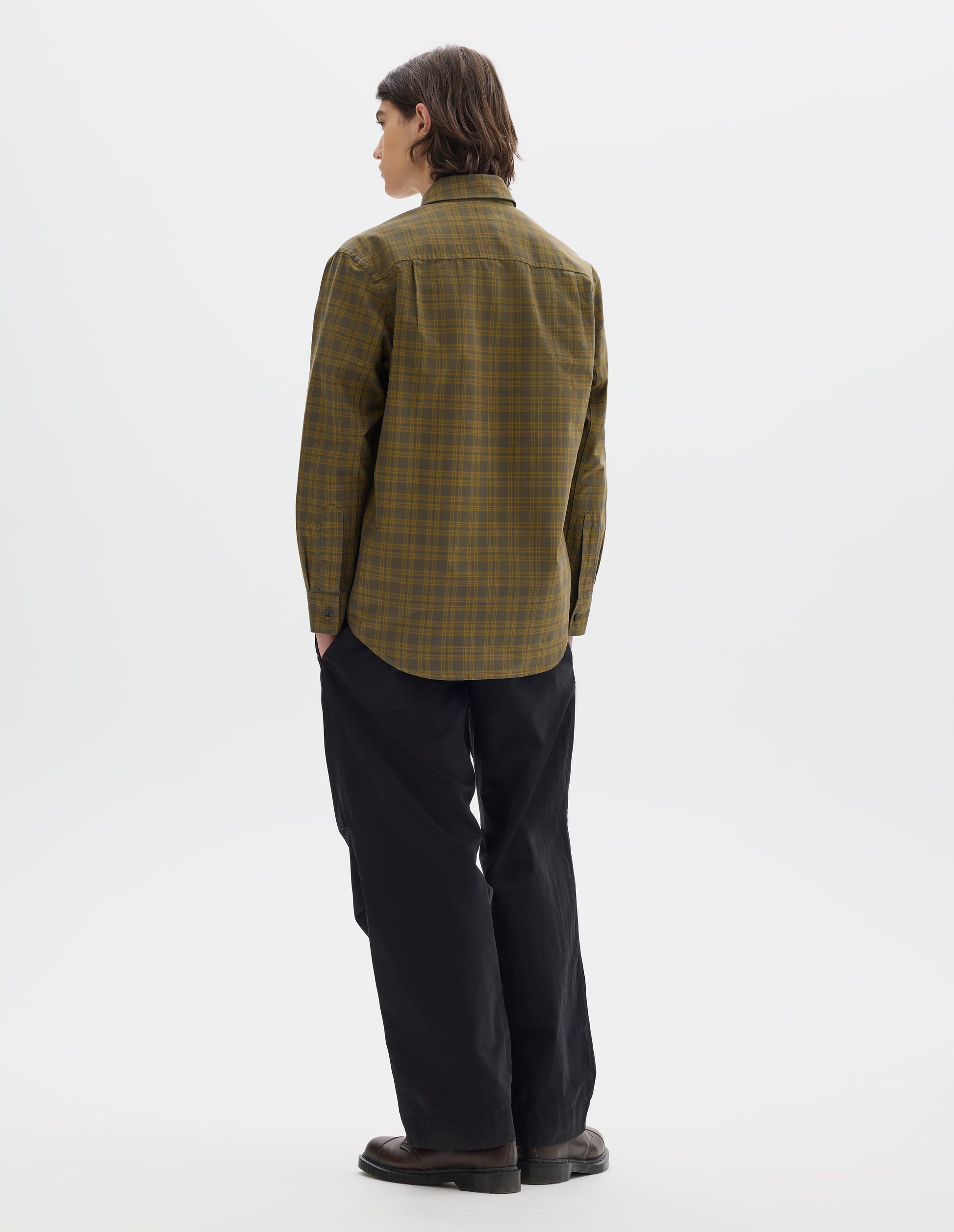 Full body image, back view of model wearing a green check shirt with a box pleat on back yoke, button-through cuff gauntlet and a curved hem. Worn with black cotton trousers and styled with brown leather shoes.