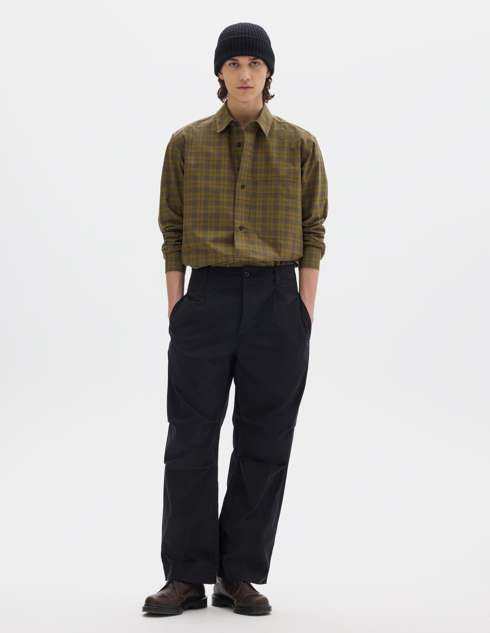 Full body image, front view of model wearing a black beanie, a green check buttoned down shirt, sleeves pulled up. Worn with black cotton trousers with side seam pockets, single front pleat and pleated knee detailing. Styled with brown leather shoes.