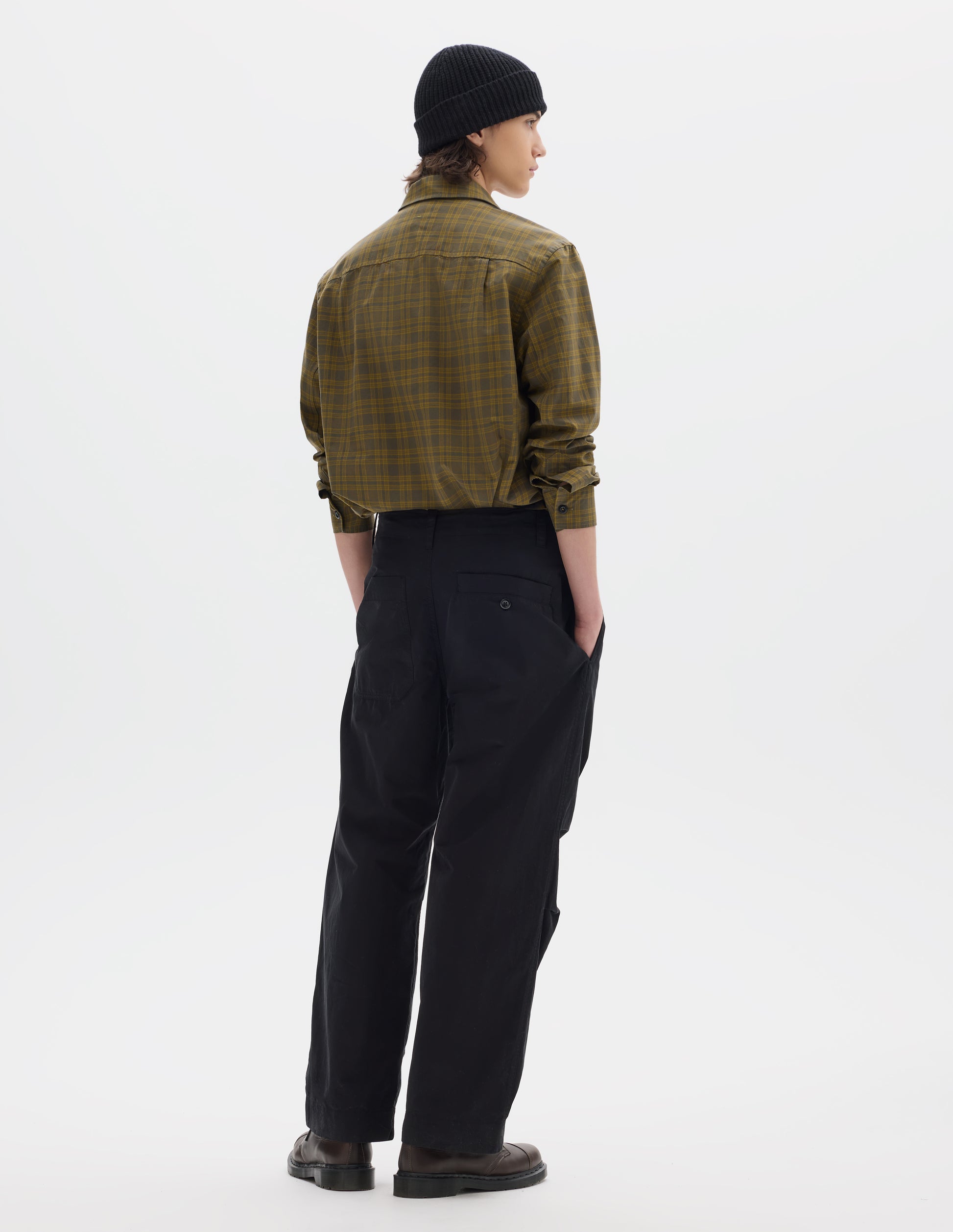 Full body image, back view of model wearing a black beanie, a green check shirt, with a stitch through yoke, sleeves pulled up. Worn with black cotton trousers with side seam pockets, two back pockets, one is a patch pocket and one a button through welt pocket. Styled with brown leather shoes.