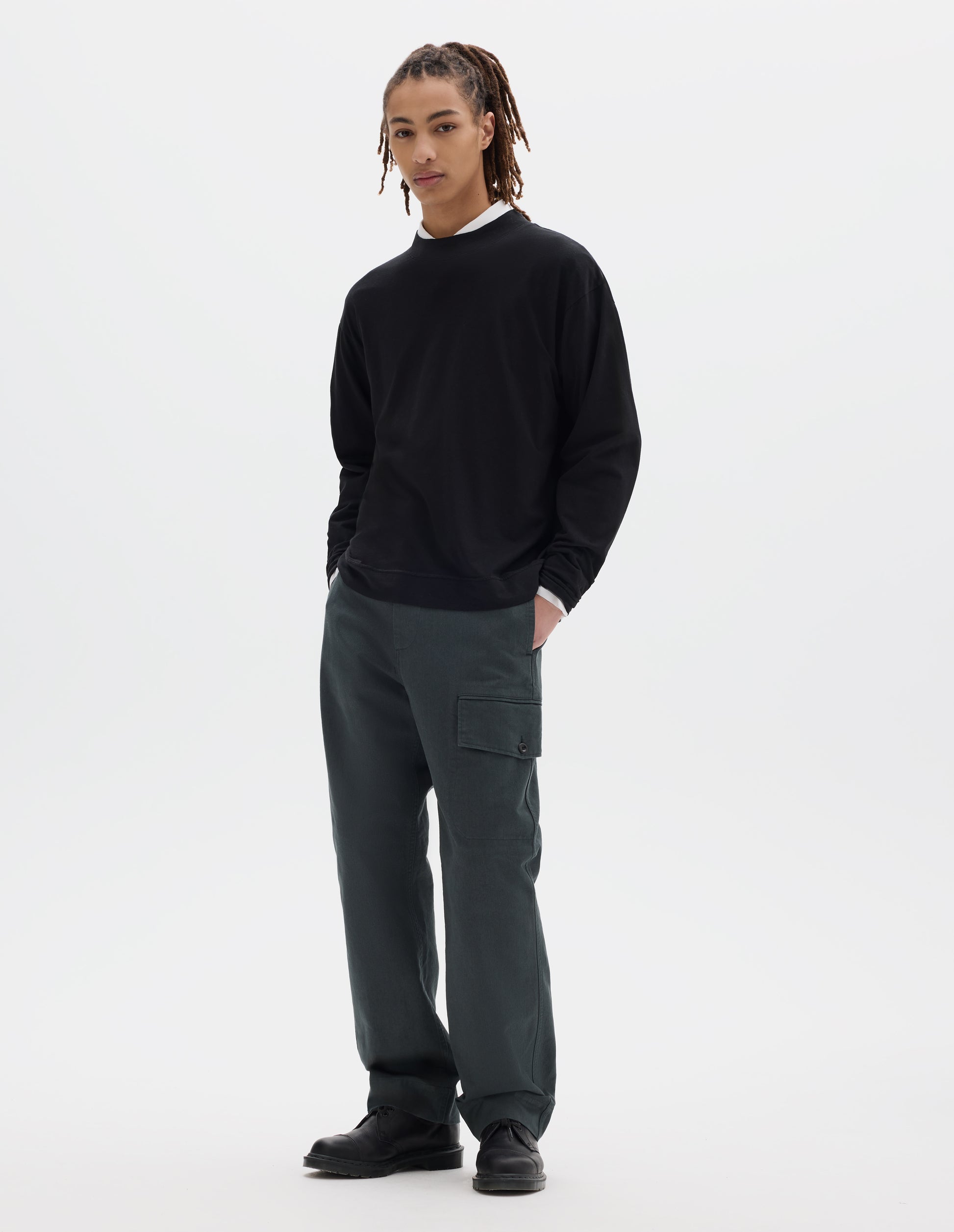 Full body image, front view of model wearing a boxy black cotton linen long sleeve crew neck jersey. Worn over a white shirt, styled with dark green-blue cargo trousers and black leather shoes.