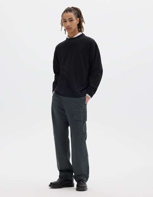 Full body image, front view of model wearing a boxy black cotton linen long sleeve crew neck jersey. Worn over a white shirt, styled with dark green-blue cargo trousers and black leather shoes.