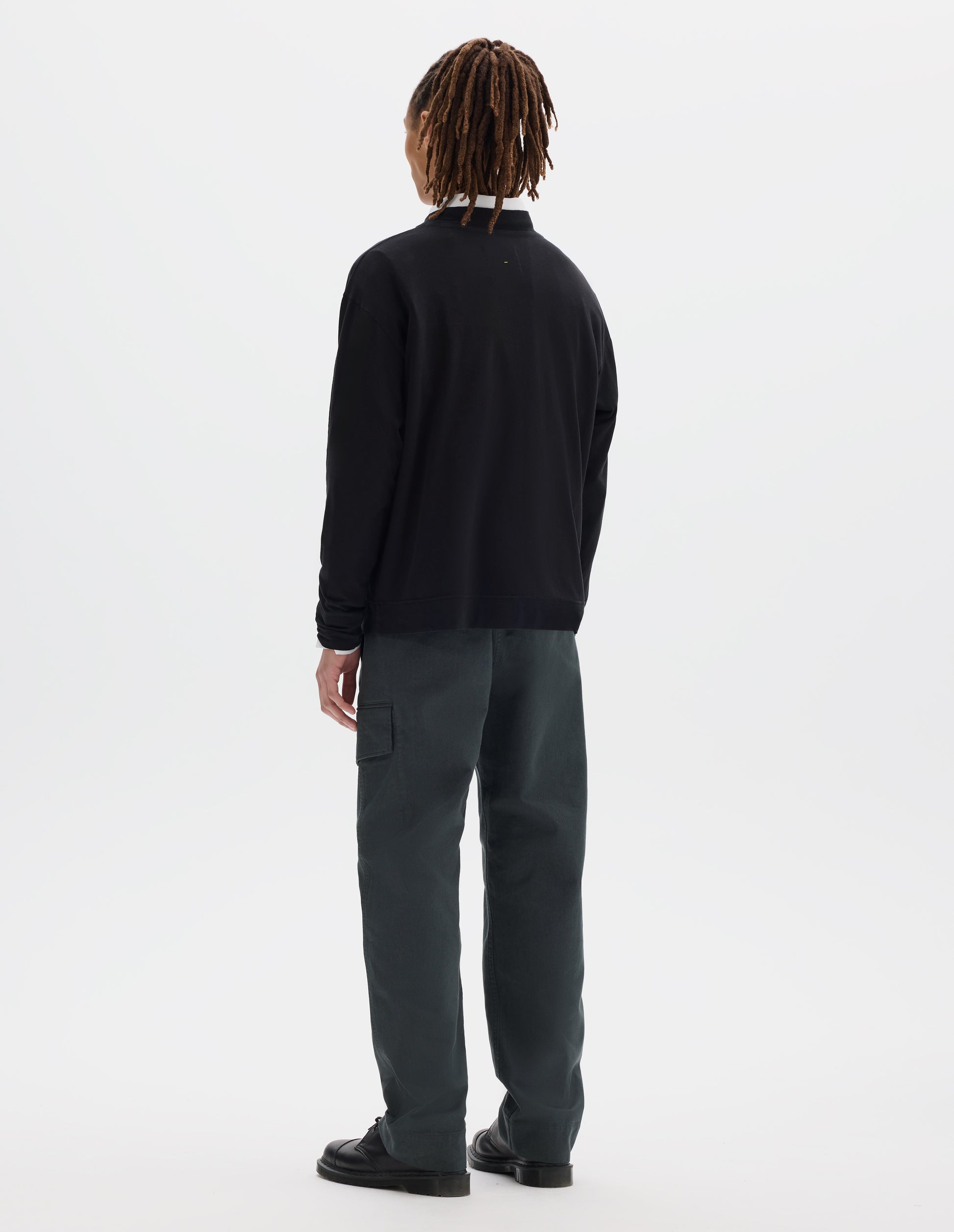 Full body image, back view of model wearing a boxy black cotton linen long sleeve crew neck jersey. Worn over a white shirt, styled with dark green-blue cargo trousers and black leather shoes.