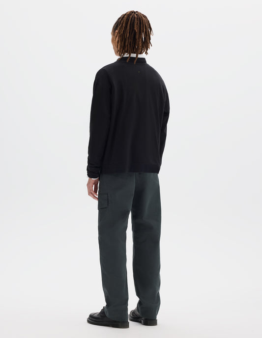 Full body image, back view of model wearing a boxy black cotton linen long sleeve crew neck jersey. Worn over a white shirt, styled with dark green-blue cargo trousers and black leather shoes.