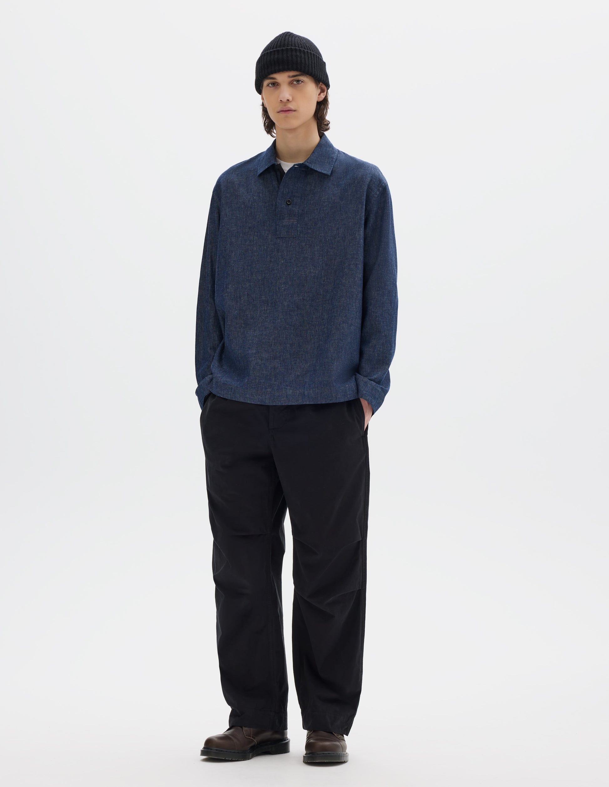 Full body image, front view of model wearing blue cotton linen polo style shirt, with a wide placket which has two dark buttons at opening. Styled over a white T shirt, black cotton trousers, brown leather shoes and a black wool beanie.