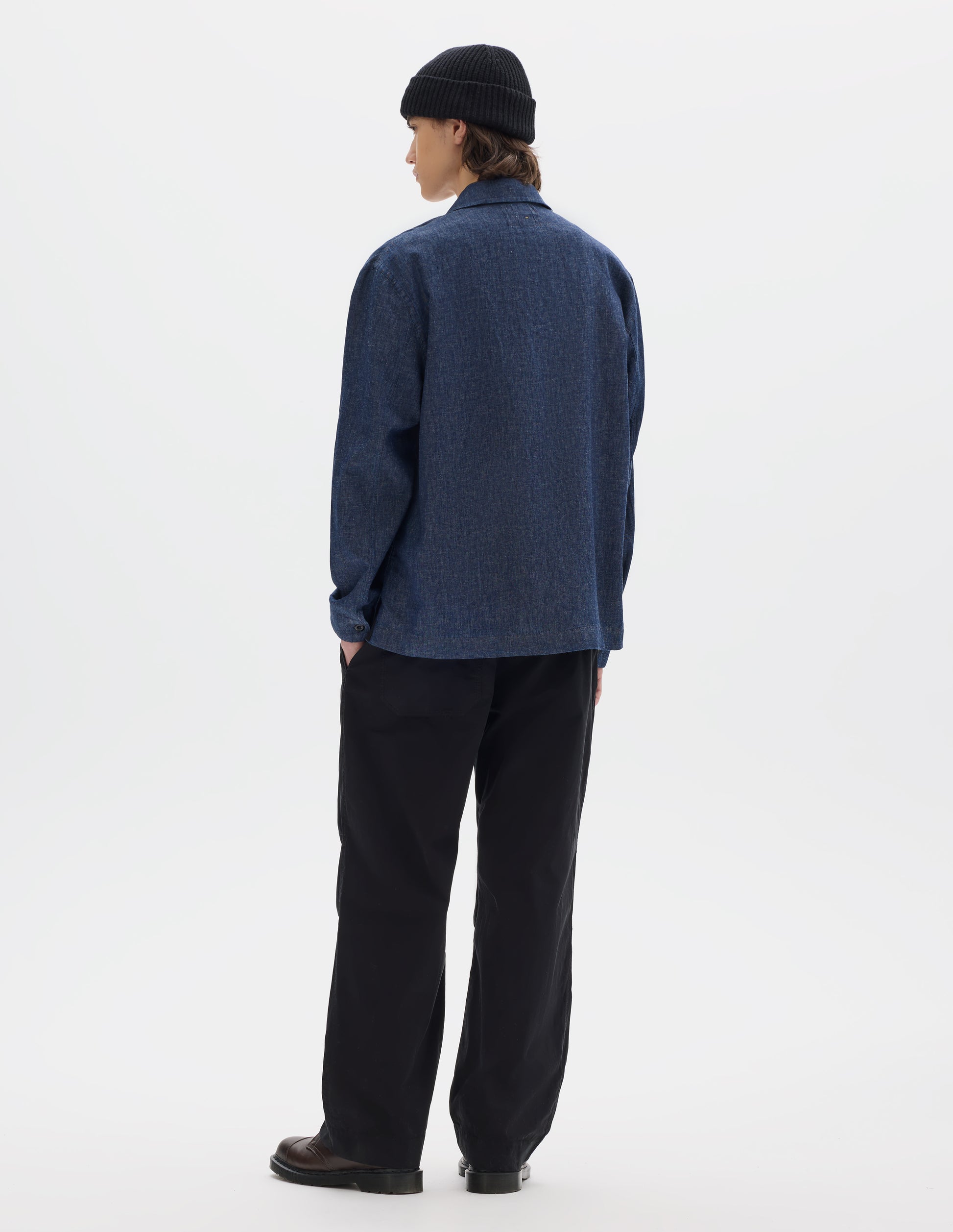 Full body image, back view of model wearing blue cotton linen polo style shirt with an extended grown-on cuffs. Styled with black trousers, brown leather shoes and a black wool beanie.