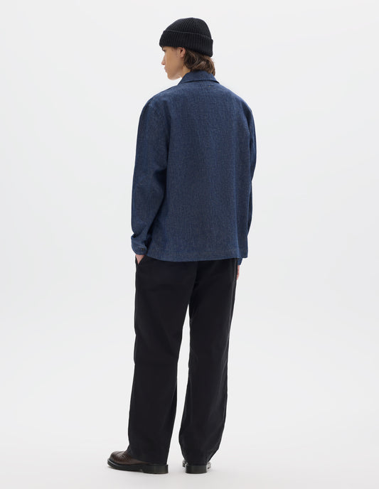 Full body image, back view of model wearing blue cotton linen polo style shirt with an extended grown-on cuffs. Styled with black trousers, brown leather shoes and a black wool beanie.