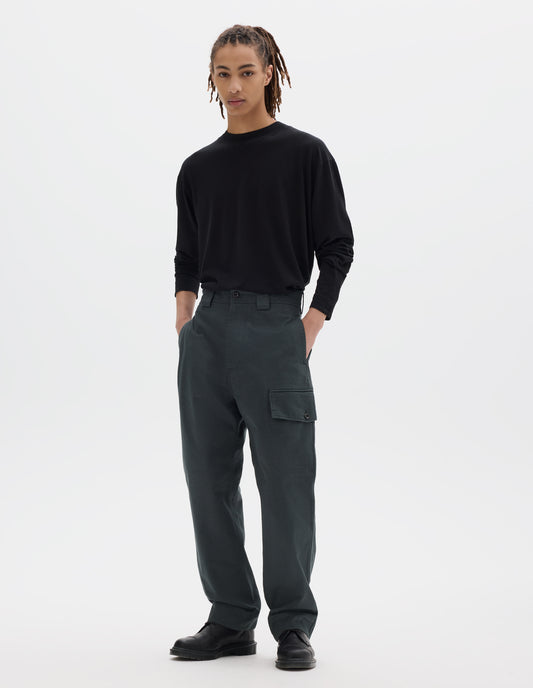 Full body image, front view of model wearing blueish grey cotton trousers, with wide belt loops and a button and zip fly fastening. With side seam pockets, a large cargo pocket with button fastening. Styled with black long sleeve tucked in and black leather shoes.
