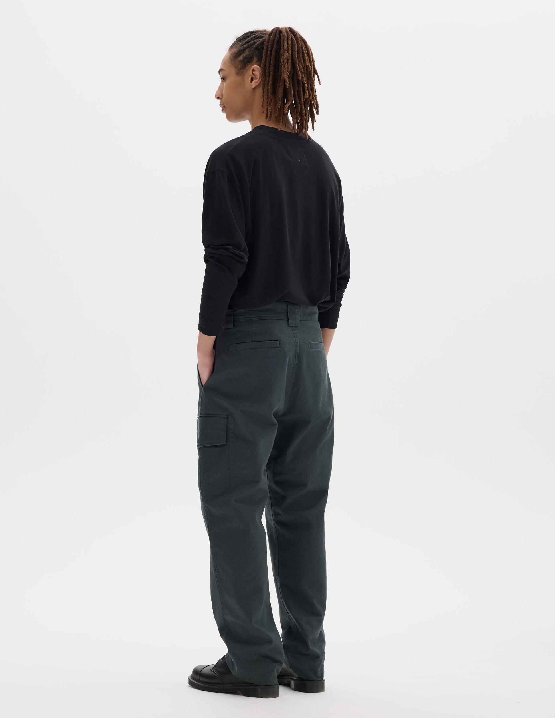 Full body image, back view of model wearing blueish grey cotton trousers, with wide belt loops, a large cargo pocket with button fastening and welt back pockets. Styled with black long sleeve tucked in and black leather shoes.