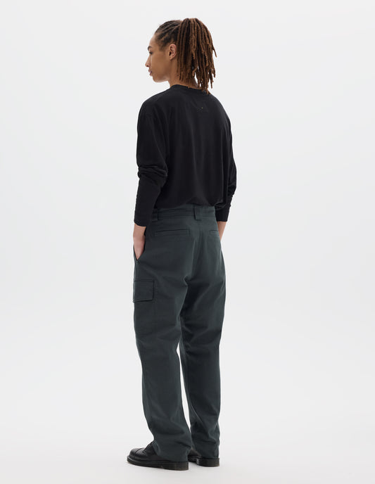 Full body image, back view of model wearing blueish grey cotton trousers, with wide belt loops, a large cargo pocket with button fastening and welt back pockets. Styled with black long sleeve tucked in and black leather shoes.