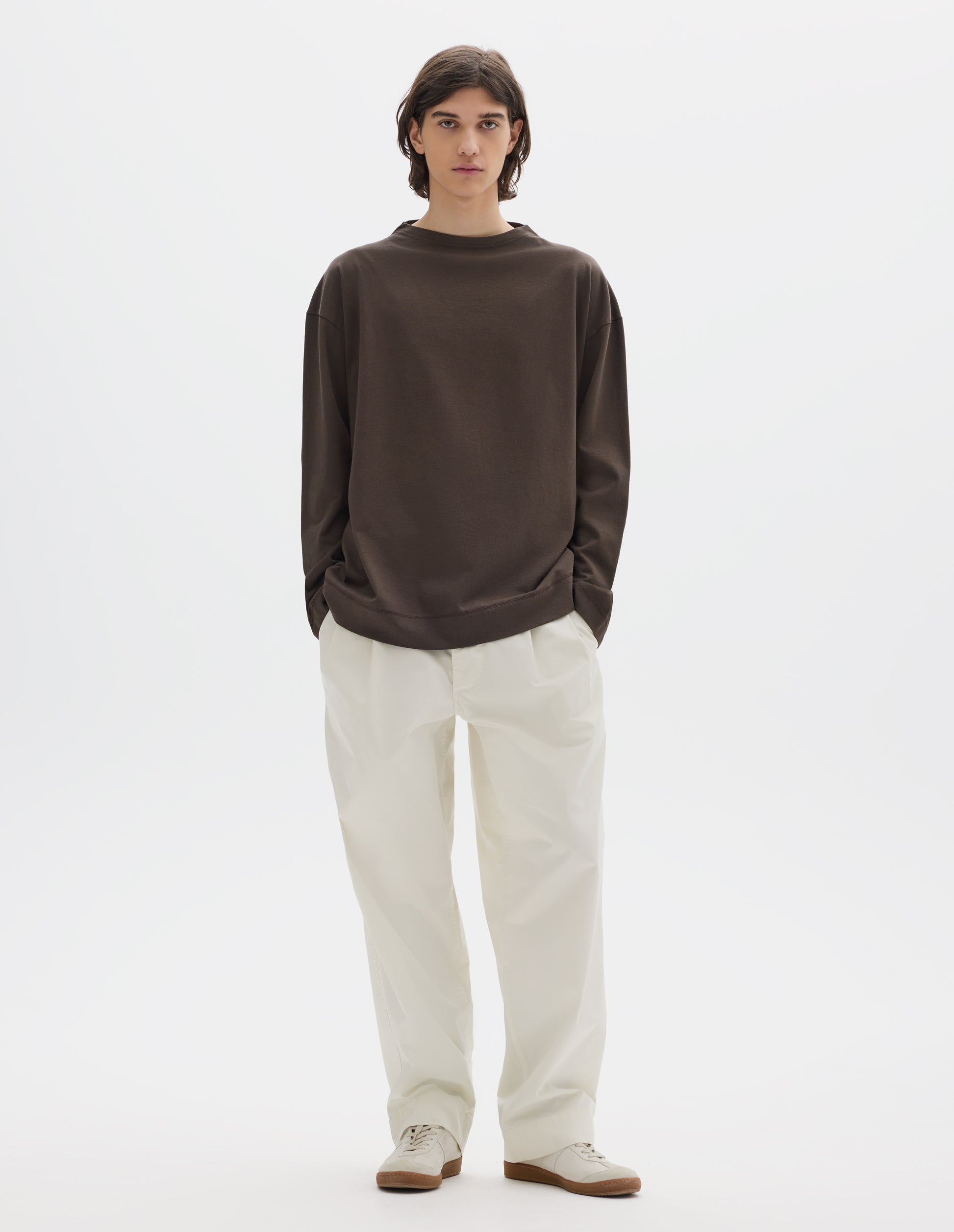 Full body image, front view of model wearing brown cotton t shirt, with long sleeves and flatlock stitching around cuffs and hem. Styled with white trousers and white shoes.