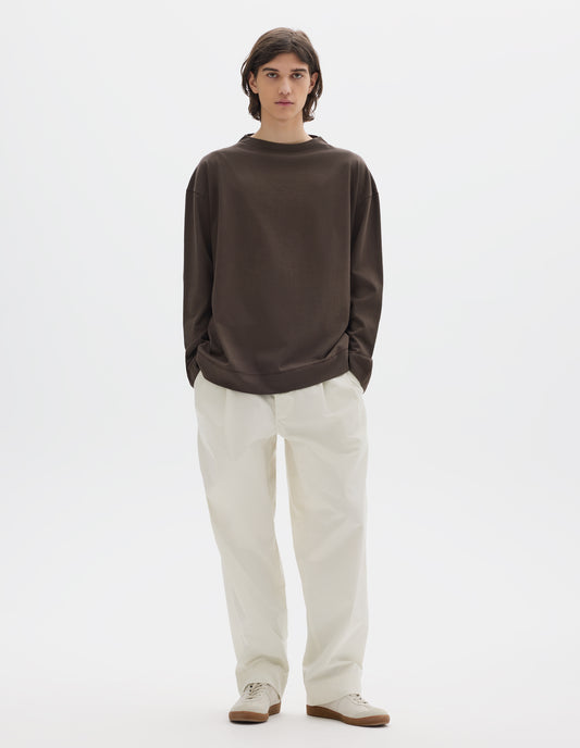 Full body image, front view of model wearing brown cotton t shirt, with long sleeves and flatlock stitching around cuffs and hem. Styled with white trousers and white shoes.