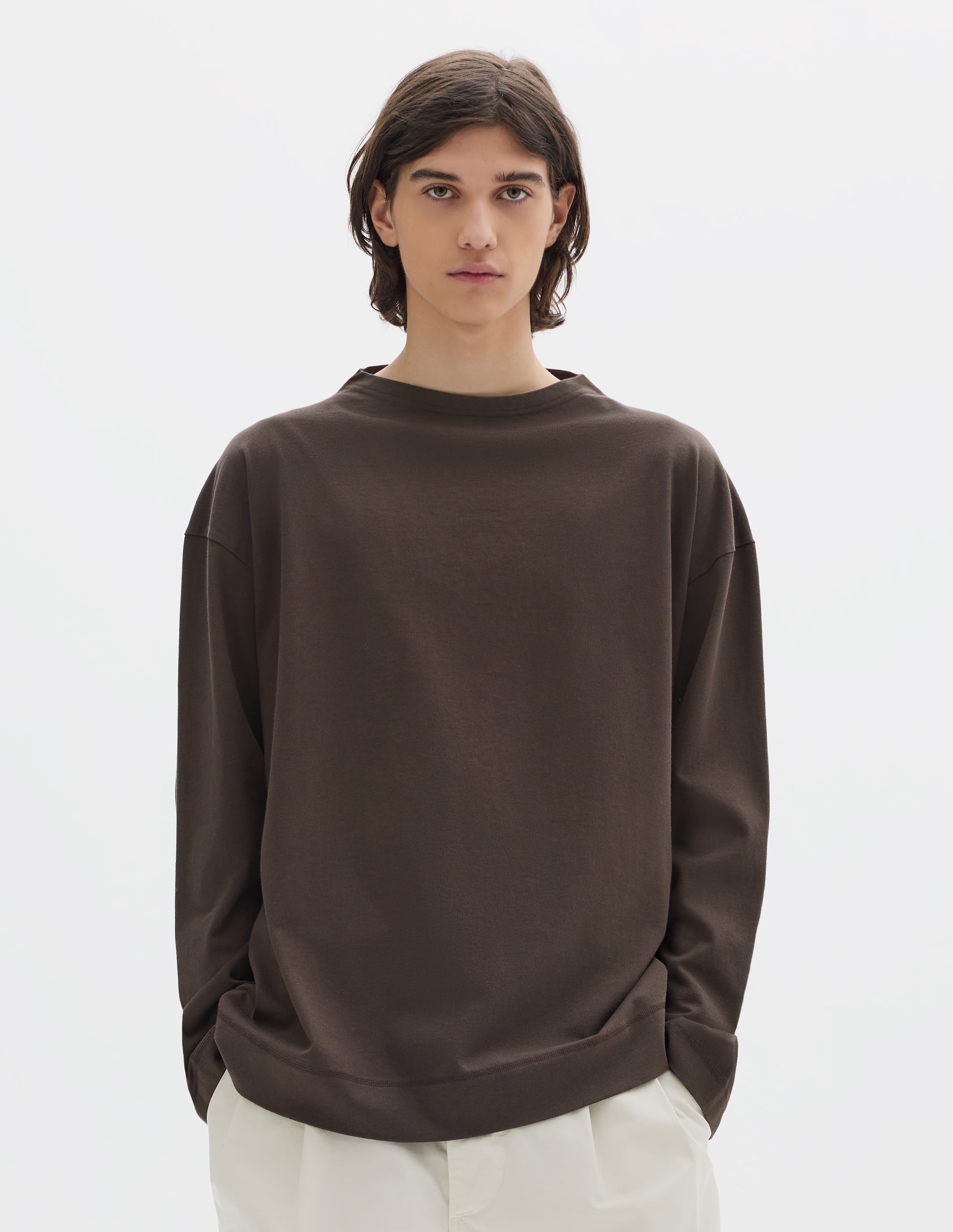 Cropped body image, front view of model wearing brown cotton t shirt, with long sleeves and flatlock stitching around cuffs and hem. Styled with white trousers.
