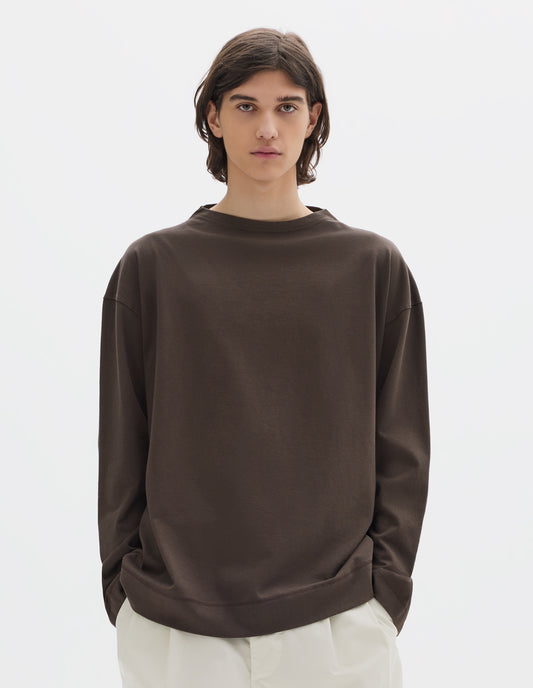 Cropped body image, front view of model wearing brown cotton t shirt, with long sleeves and flatlock stitching around cuffs and hem. Styled with white trousers.