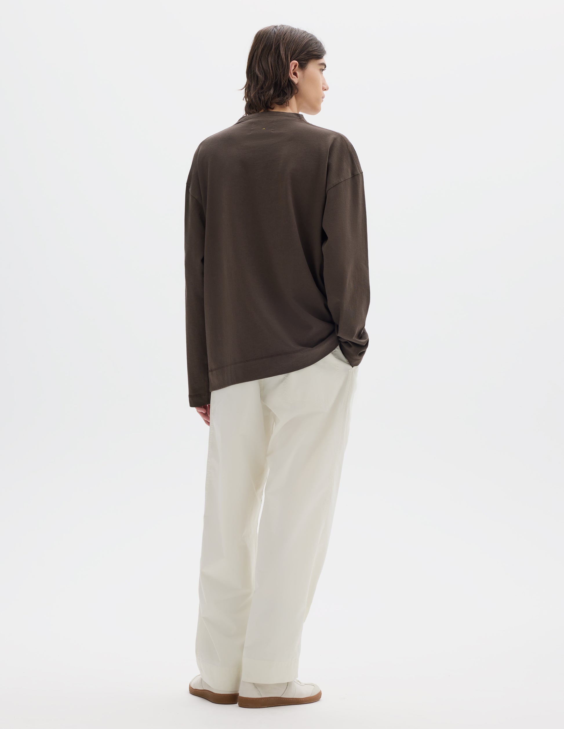 Full body image, back view of model wearing brown cotton t shirt, with long sleeves and flatlock stitching around cuffs and hem. Styled with white trousers and white shoes.