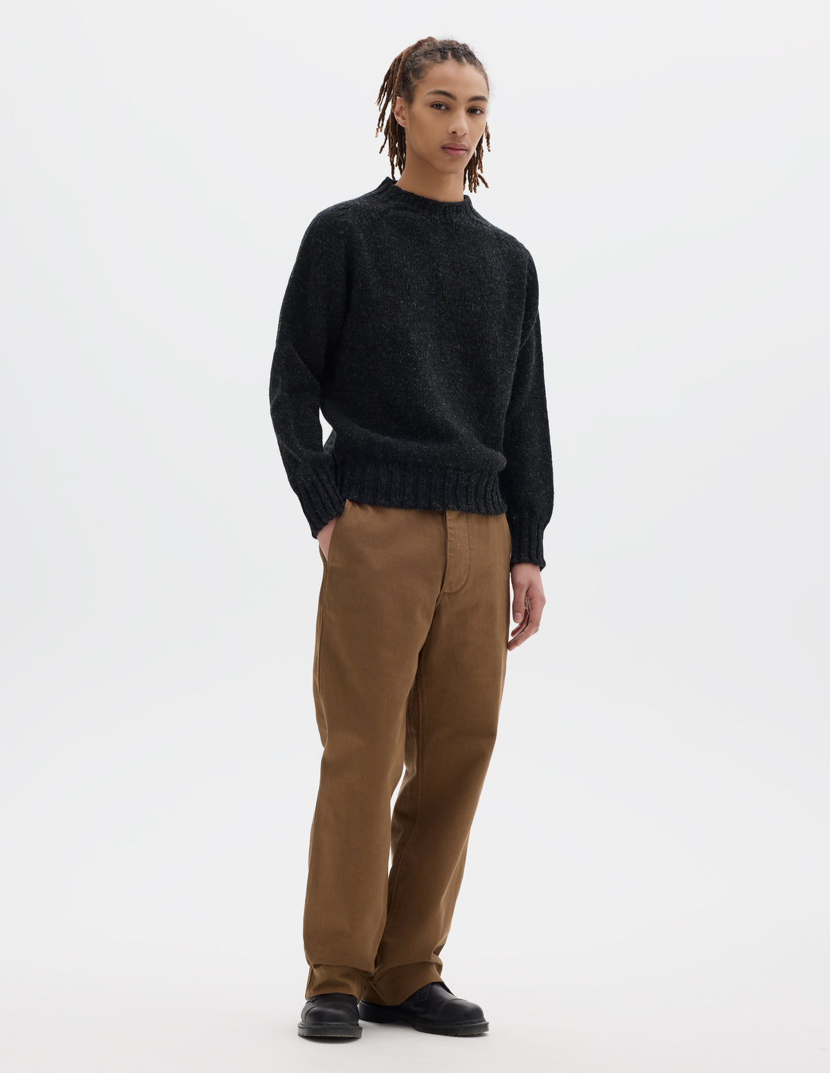 Full body image, front view of model wearing dark grey long sleeve wool jumper, with contrast ribbing on neckline, cuffs and hem. Styled with brown trousers and black leather shoes.