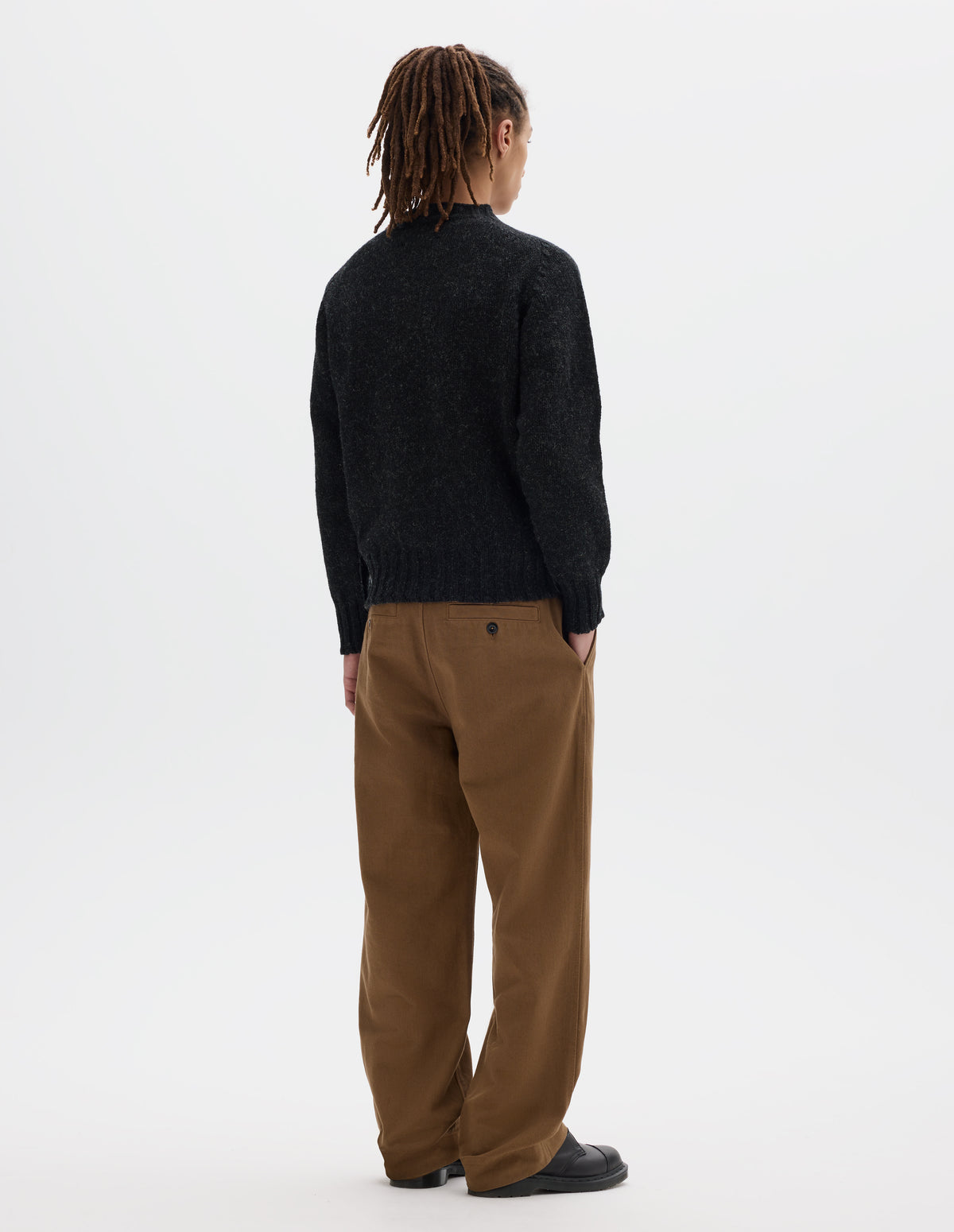 Full body image, back view of model wearing dark grey long sleeve wool jumper, with contrast ribbing on neckline, cuffs and hem. Styled with brown trousers and black leather shoes.