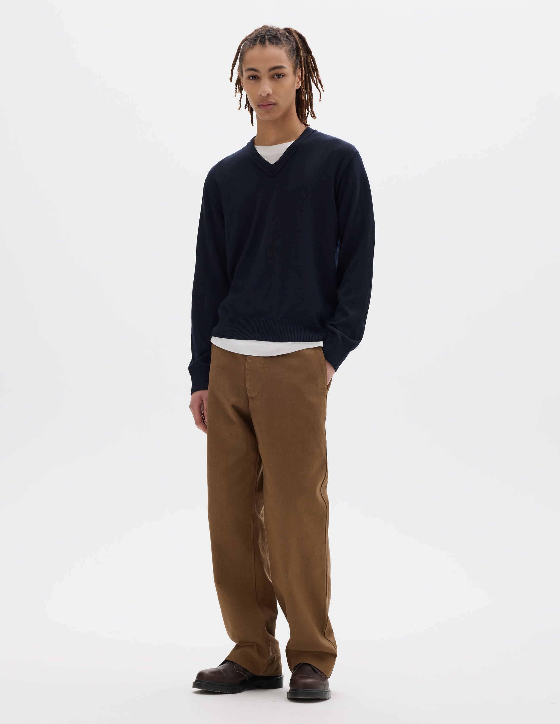 Full body image, front view of model wearing dark navy fine wool jumper with v neck, finished with rib on cuffs and hem. Styled over a white t shirt, brown trousers and dark brown leather shoes.