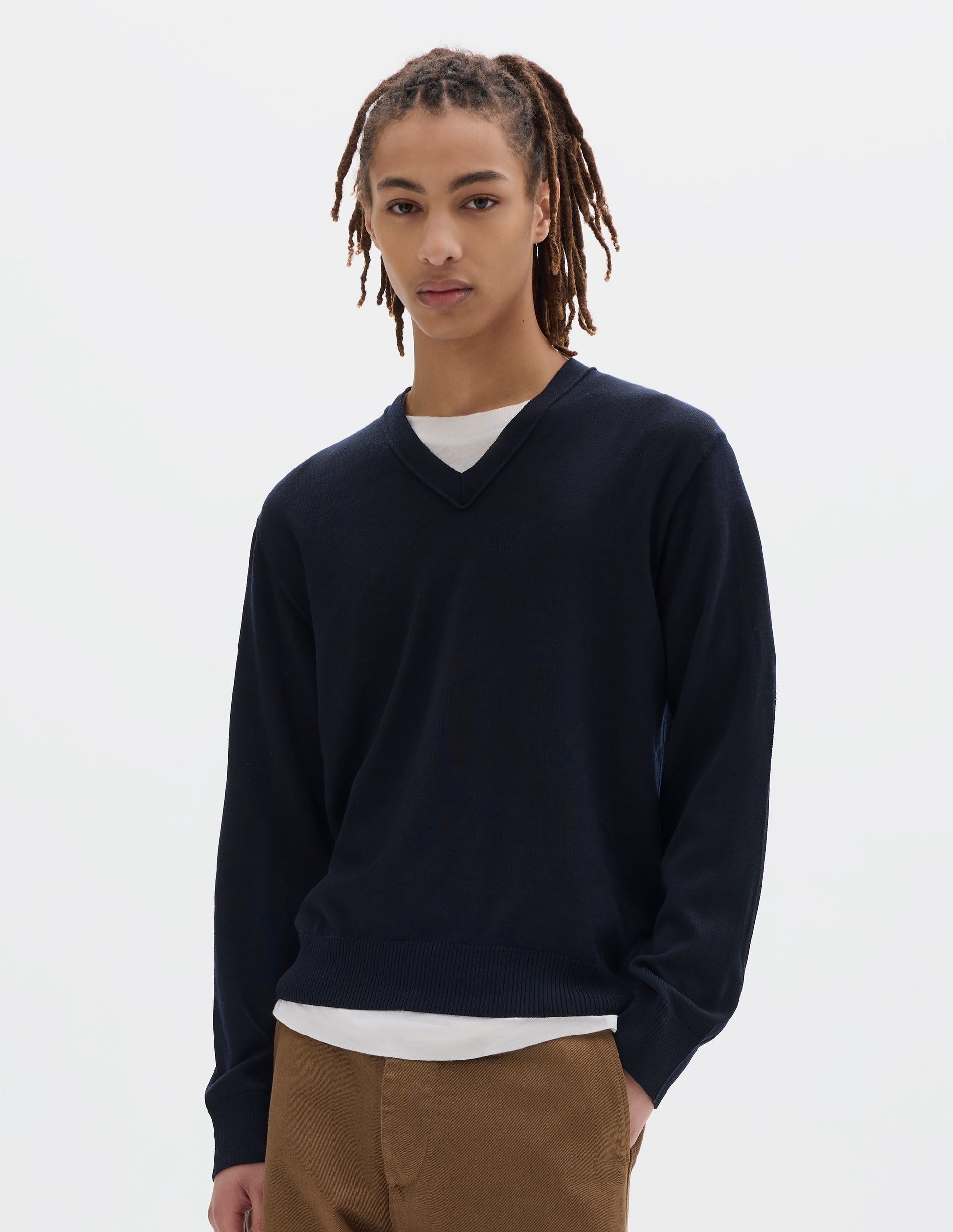 Cropped body image, front view of model wearing dark navy fine wool jumper with v neck, finished with rib on cuffs and hem. Styled over a white t shirt and brown trousers.