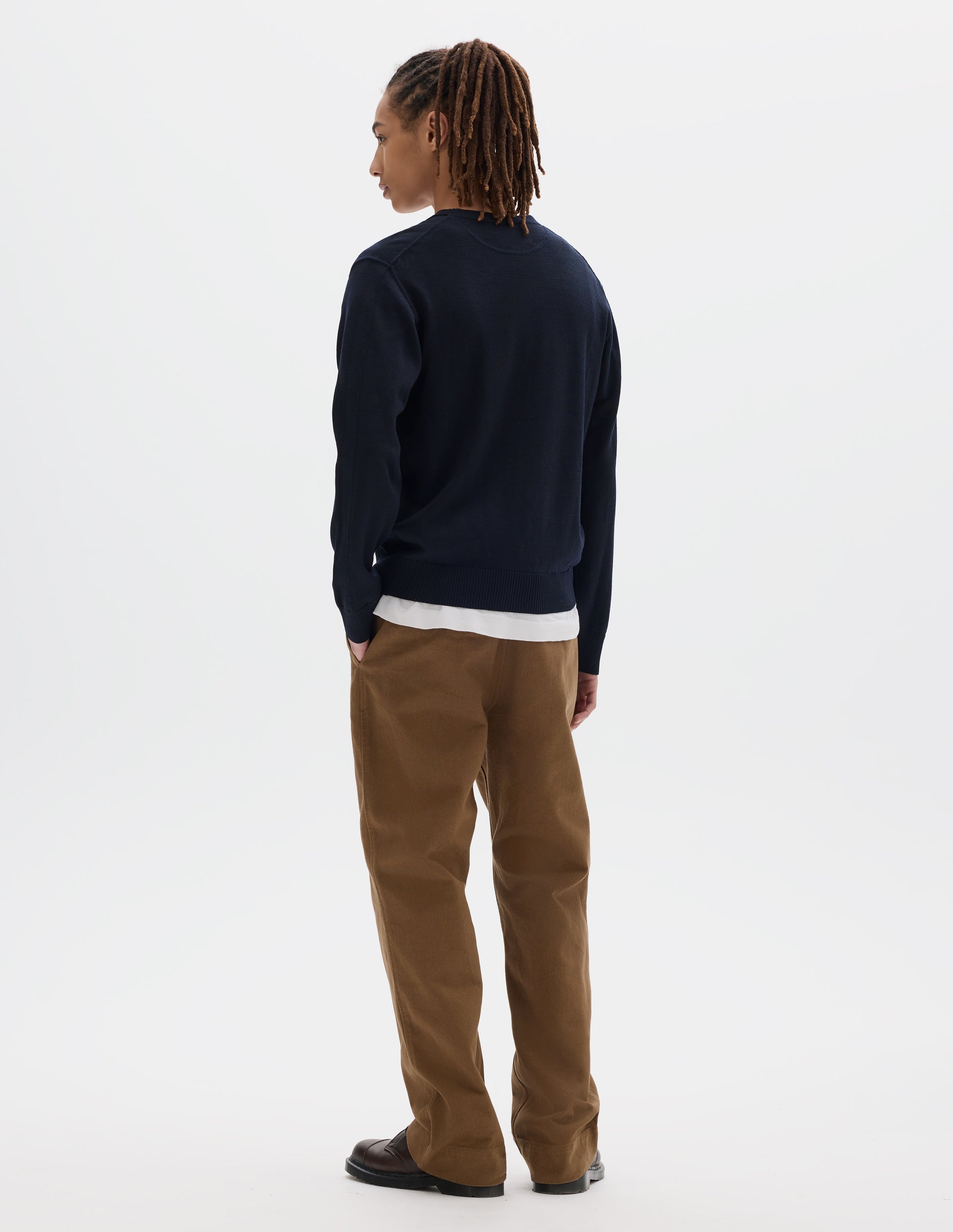 Full body image, back view of model wearing dark navy fine wool jumper with rib on cuffs and hem. Styled over a white t shirt, brown trousers and dark brown leather shoes.